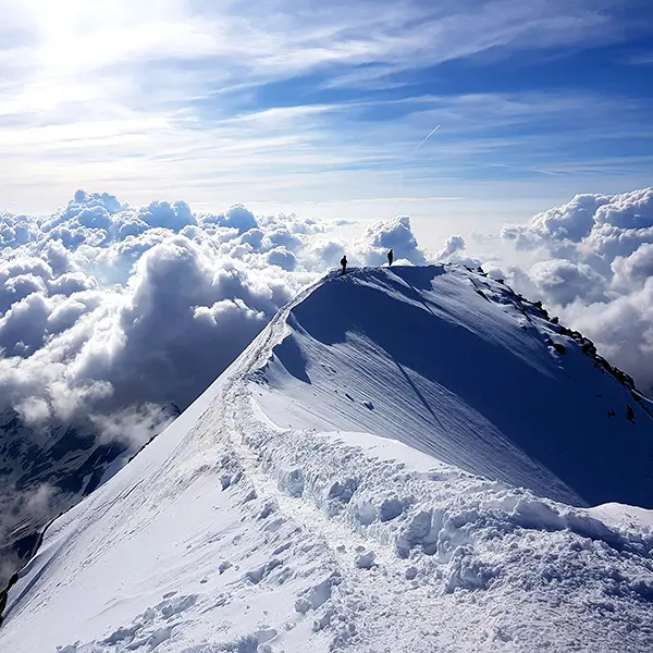 Twee bergbeklimmers staan op een besneeuwde bergtop boven de wolken, met een indrukwekkende lucht en zonlicht dat het wi